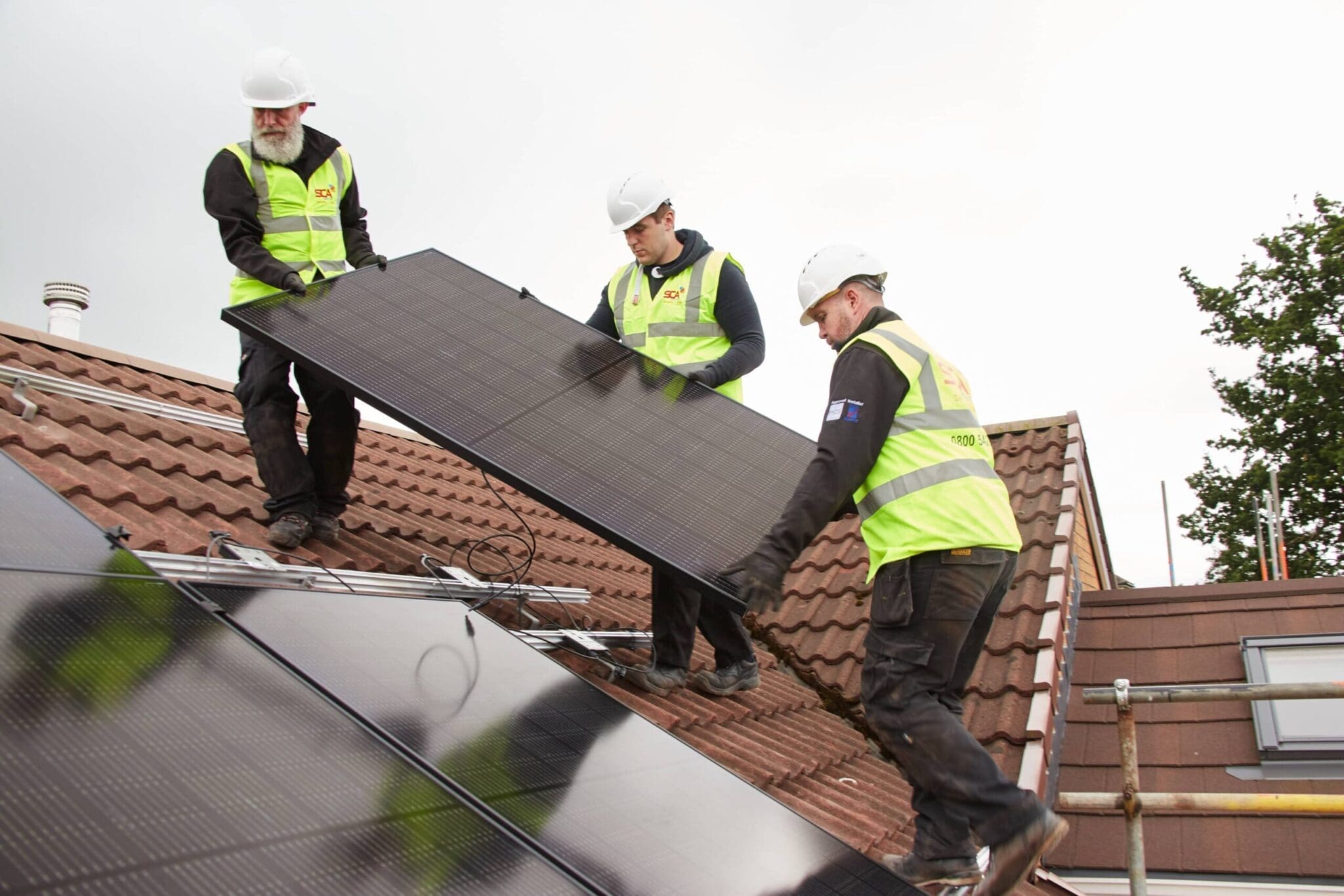 Three workers in safety gear install solar panels on a home roof.